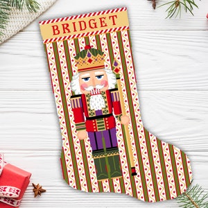 May include: A festive Christmas stocking with a pixelated nutcracker design. The stocking has a cream-coloured top with the name "BRIGET" in red. The body of the stocking features a green and red striped pattern with a nutcracker in the centre, holding a staff.