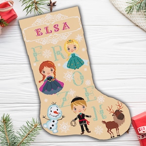 Op de afbeelding: Een beige kerstsok met de naam "Elsa" geborduurd in roze letters. De sok heeft een sneeuwvlokkenpatroon en personages uit de film Frozen, waaronder Elsa, Anna, Olaf en Kristoff.
