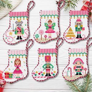 Op de afbeelding: Zes roze en witte kerstsokken met een Notenkraker-thema. Elke sok heeft een ander personage uit het Notenkraker-verhaal, waaronder de Notenkraker, de Suikerfee en de Muizenkoning. De sokken zijn versierd met snoepstokken, sneeuwvlokken en sterren.