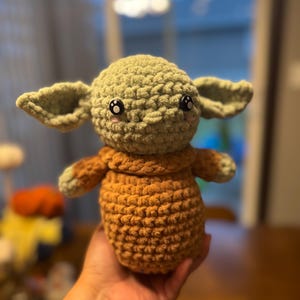May include: A crocheted plush toy resembling a character from the Star Wars franchise. The toy is green with large eyes and a brown crocheted body.