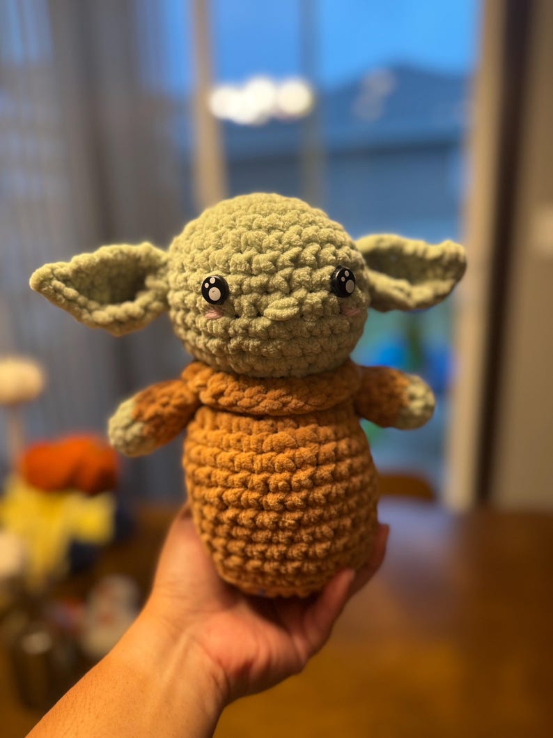 May include: A crocheted plush toy resembling a character from the Star Wars franchise. The toy is green with large eyes and a brown crocheted body.