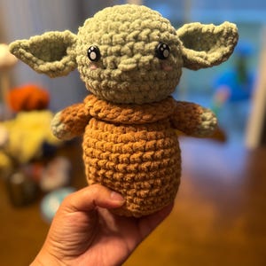May include: A crocheted plush toy in the shape of a green alien character with large ears, brown arms and legs, and black eyes.