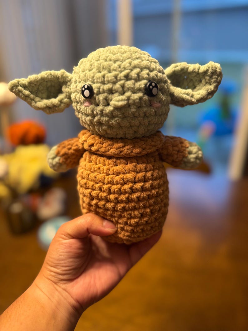 May include: A crocheted plush toy in the shape of a green alien character with large ears, brown arms and legs, and black eyes.