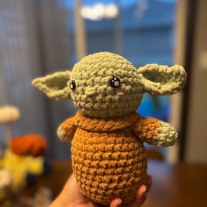 May include: A crocheted plush toy resembling a character from the Star Wars franchise. The toy is green with a brown sweater and has large black eyes with white pupils.