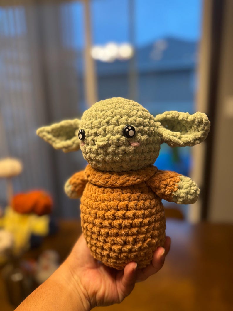 May include: A crocheted plush toy resembling a character from the Star Wars franchise. The toy is green with a brown sweater and has large black eyes with white pupils.