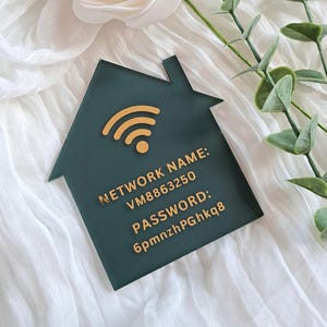 May include: A house-shaped dark green sign featuring a gold Wi-Fi symbol and the text: NETWORK NAME: VM8863250 PASSWORD: 6pmnzhPGhkq8. The sign is displayed against a white fabric background with a white rose and green leaves.