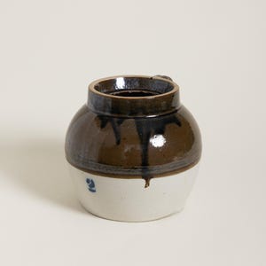 May include: A small, round, ceramic jar with a dark brown glazed top and a white base. The jar has a small handle and a blue number 2 painted on the white base. The glaze drips down the side.
