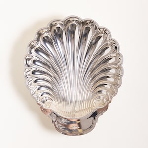 May include: A silver-colored, shell-shaped serving dish. The dish has a scalloped edge and a detailed, ribbed interior. The base of the dish has a decorative, curved design. The dish is made of a shiny, reflective metal.