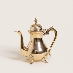 May include: A polished gold-colored teapot with a curved spout and handle. The teapot has a domed lid with a finial and rests on three small feet. The design includes decorative shell-like accents near the base.