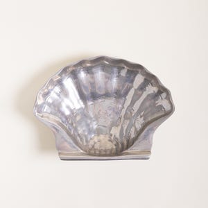 May include: A silver-colored, shell-shaped decorative dish. The dish has a scalloped edge and a textured surface, with a small base for stability. The dish is made of a shiny, reflective material.