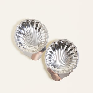 May include: Two silver-toned metal dishes shaped like scallop shells. Each dish features a polished, reflective surface with deep, radiating fluted grooves that extend from the base to the scalloped, curved edges of the shell-like form.