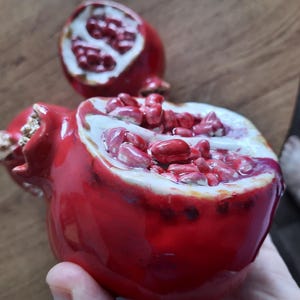 May include: Two ceramic pomegranate sculptures with a glossy red finish. One piece is halved to reveal detailed, textured red seeds against a white interior, while the other is partially open, showing a similar red and white seed structure.