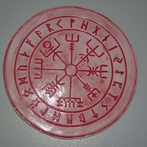 May include: A circular, red decorative piece with a central symbol and surrounding runes. The design resembles a compass or emblem, possibly of historical or cultural origin. The item has a textured surface. The diameter is approximately 15 cm.