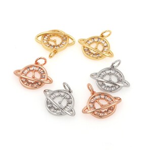 May include: Five gold, silver, and rose gold charm pendants in the shape of a planet with a ring around it. The planet is decorated with small, clear rhinestones.
