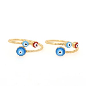 Könnte beinhalten: Zwei goldene Ringe mit blauen und roten bösen Augen-Anhängern. Die Ringe sind verdreht und haben eine strukturierte Oberfläche.