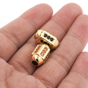 May include: Two gold-toned metal beads with a square shape and a rounded end. One bead is decorated with black rhinestones and the other with orange rhinestones.