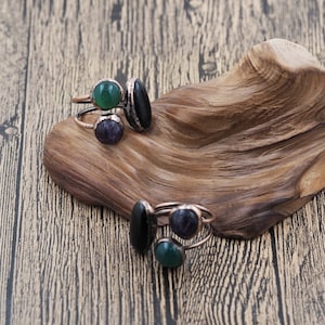 May include: Four copper-colored rings with various gemstone settings. The gemstones are green, black, and purple. The rings are displayed on a wooden, organic-shaped display.