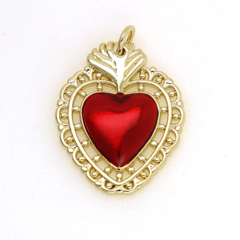 May include: A gold-tone pendant featuring a red heart with a flame design at the top. The heart is surrounded by a delicate filigree frame.