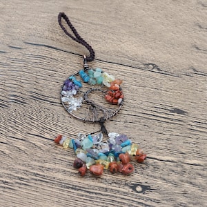 May include: A wire-wrapped tree of life charm featuring an assortment of colourful gemstones, including clear, red, orange, yellow, green, blue, and purple. The charm is attached to a black cord and hangs from a wooden surface.