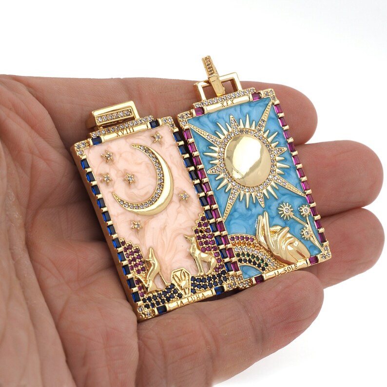May include: Two gold-tone pendants with enamel and crystal accents. The pendant on the left features a crescent moon and stars on a pink background with the text "La Luna". The pendant on the right features a sun on a blue background with the text "Il Sole".