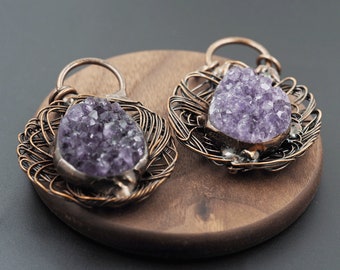 May include: Two amethyst pendants with a wire-wrapped design. Each pendant features a cluster of purple crystals set in a metal frame, surrounded by intricate copper-colored wirework. The pendants are displayed on a wooden surface.