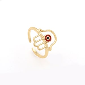 Pu&ograve; includere: Anello in oro con un design a mano di hamsa e un occhio turco rosso al centro.