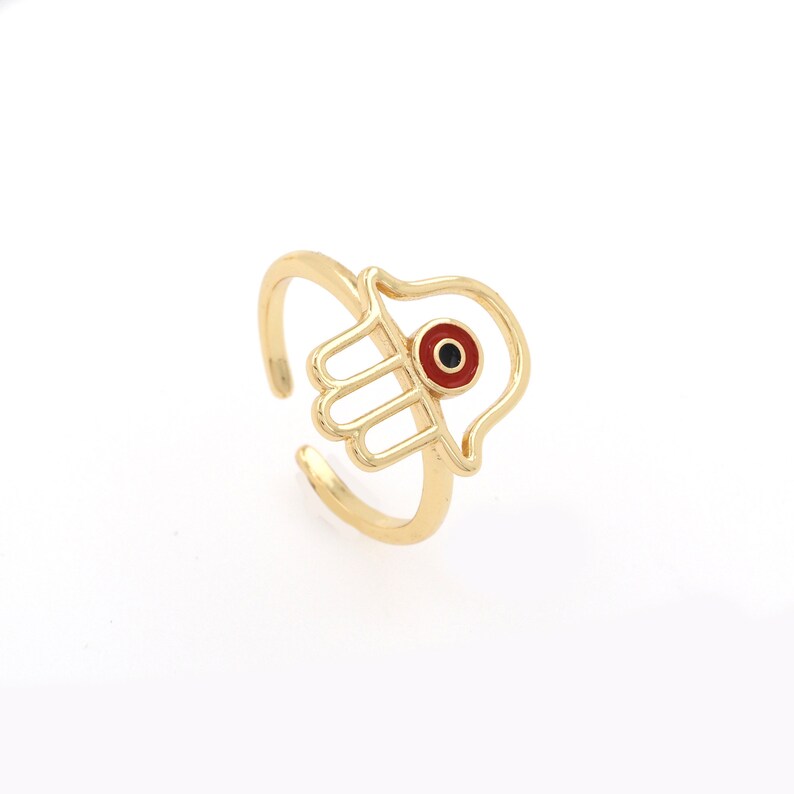 Pu&ograve; includere: Anello in oro con un design a mano di hamsa e un occhio turco rosso al centro.