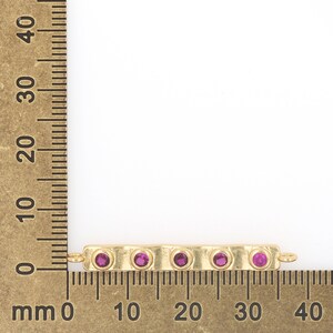 May include: Gold bar charm with five pink gemstones, measured on a ruler in inches.