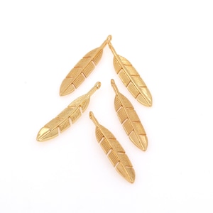 May include: Five gold-coloured feather charms with a detailed vein design. Each charm has a small loop at the top for attaching to jewellery. The feathers are arranged on a white background.