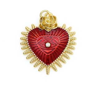 May include: A red heart-shaped pendant with a gold-colored eye and a gold-colored flower on top. The heart is surrounded by a halo of gold-colored spikes.