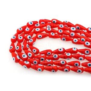 Op de afbeelding: Close-up van een streng rode glazen kralen met een druppelvorm. Elke kraal heeft een wit oog met een blauwe iris. De kralen zijn geregen, waardoor een decoratief element ontstaat. De achtergrond is wit.