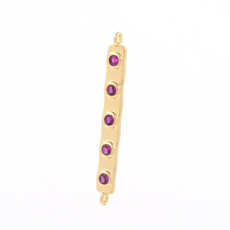 May include: Gold bar connector with five purple gemstones. The connector has two loops at the top and bottom.