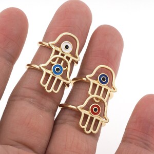Pu&ograve; includere: Tre anelli d'oro con un design a mano di hamsa. Ogni anello presenta un ciondolo di occhio turco di colore diverso: blu, bianco e rosso.