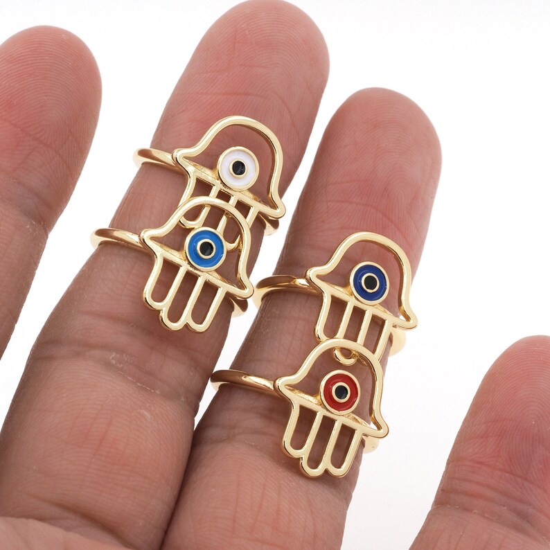 Pu&ograve; includere: Tre anelli d'oro con un design a mano di hamsa. Ogni anello presenta un ciondolo di occhio turco di colore diverso: blu, bianco e rosso.