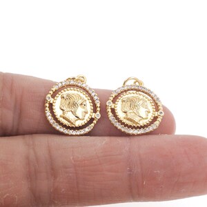 May include: Two gold-toned pendant charms featuring a profile of a person's face surrounded by a circle of clear rhinestones.