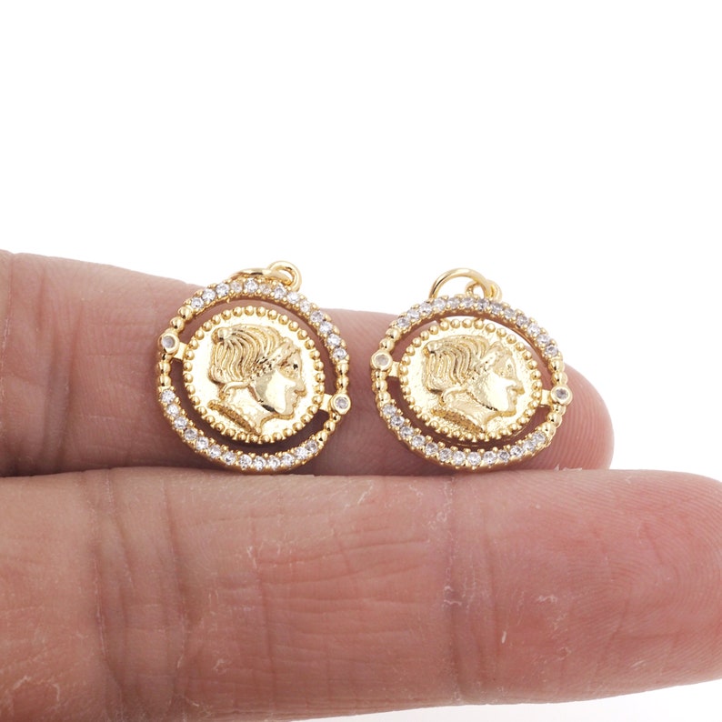 May include: Two gold-toned pendant charms featuring a profile of a person's face surrounded by a circle of clear rhinestones.