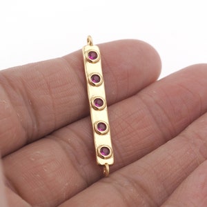 May include: Gold bar pendant with five round, purple gemstones. The pendant has two loops at the top for attaching to a chain.