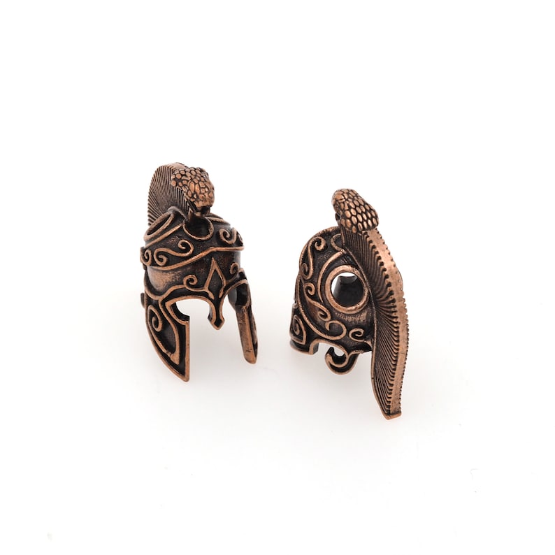 May include: Two ornate copper-colored helmet charms with intricate designs. The helmets have a raised crest and a snake design on the top.