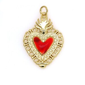 May include: A gold-colored heart-shaped pendant with a red enamel heart in the center. The pendant has a detailed design with a decorative border and a flame-like top.