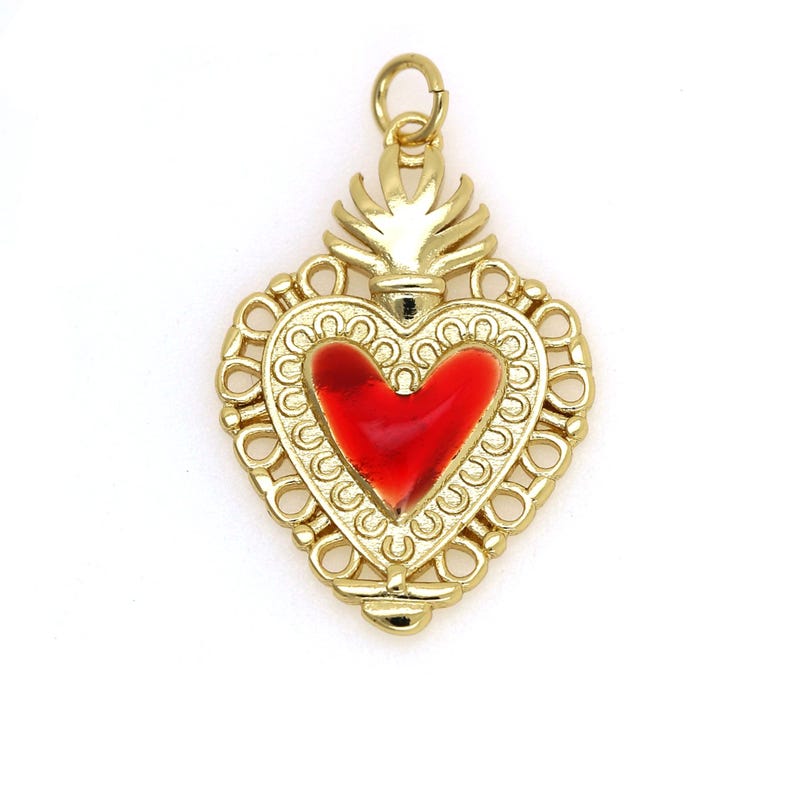 May include: A gold-colored heart-shaped pendant with a red enamel heart in the center. The pendant has a detailed design with a decorative border and a flame-like top.