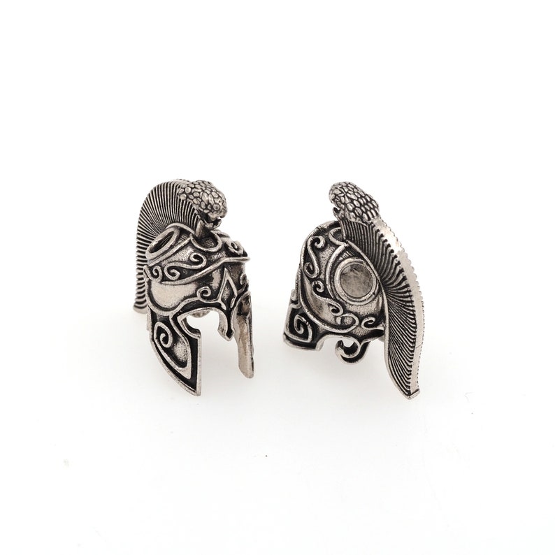 May include: Two silver-toned, detailed, miniature Roman helmet charms. Each helmet has a plume on the top and intricate designs on the sides.