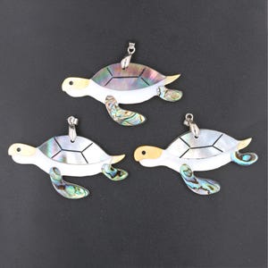 Könnte beinhalten: Drei schillernde Meeresschildkröten-Anhänger mit silberfarbenen Metallscharnieren. Die Schildkröten sind aus Perlmutt gefertigt und haben einen weißen Hintergrund mit schillernden grünen, blauen und violetten Akzenten.