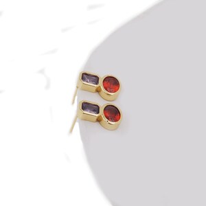 May include: A pair of gold earrings with a purple and red gemstone design. The earrings have a small, round red gemstone and a rectangular purple gemstone.