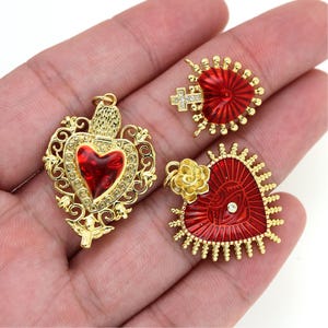 May include: Three gold-tone heart-shaped pendants with red enamel and rhinestones. The pendants feature intricate designs and are perfect for jewelry making.