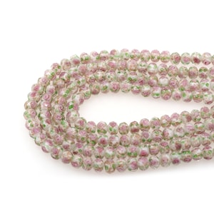 Puede incluir: Una hebra de cuentas de vidrio blanco con un patrón floral rosa y verde. Las cuentas son facetadas y tienen un acabado ligeramente iridiscente.
