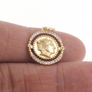 May include: Gold-toned pendant featuring a round coin with a profile of a woman's face. The coin is surrounded by a circle of small clear stones.
