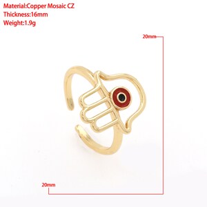 Pu&ograve; includere: Anello dorato con un design a mano di Hamsa e un ciondolo di occhio turco rosso. L'anello ha un diametro di 20 mm.