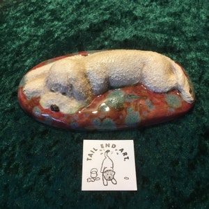 May include: A ceramic sculpture of a white dog lying down on a red, green and brown oval base. The sculpture is titled "Tail End Art".