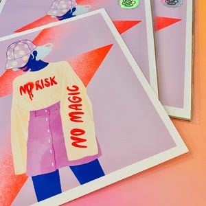 May include: A square art print featuring a stylized illustration of a figure wearing a polka-dot cap, a cream long-sleeve shirt with the red text NO RISK NO MAGIC, and a pink button-front skirt. A large orange star is in the background.