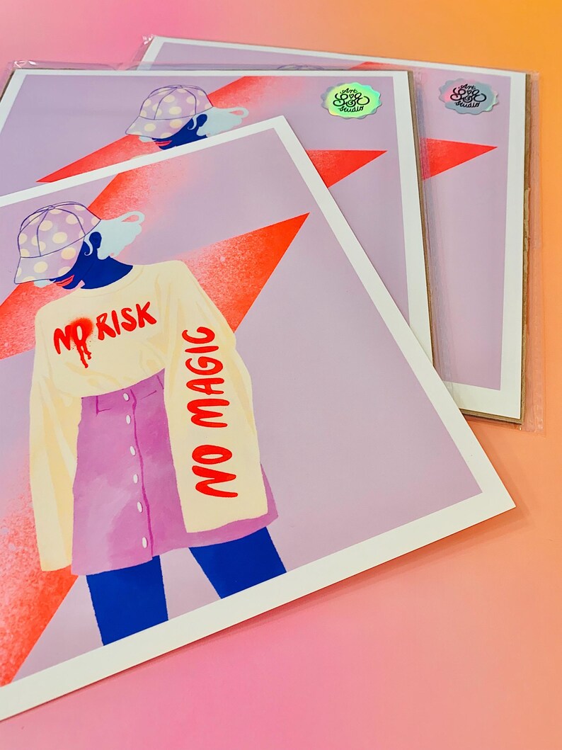 May include: A square art print featuring a stylized illustration of a figure wearing a polka-dot cap, a cream long-sleeve shirt with the red text NO RISK NO MAGIC, and a pink button-front skirt. A large orange star is in the background.